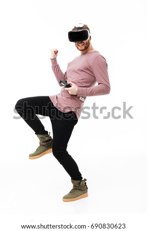 Young man standing in virtual reality glasses with joystick in hand and showing "yes" gesture on white background. Cool boy playing in visual reality glasses isolated