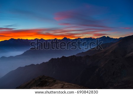 Colorful sunlight behind majestic mountain peaks of the Italian - French Alps, viewed from distant. Fog and mist covering the valleys below, autumnal landscape, cold feeling. Royalty-Free Stock Photo #690842830