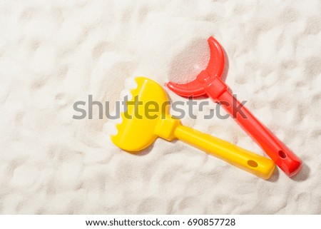 Close-up of red shovel and yellow rake on sand.