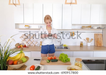 Charming woman using mobile gadget records video of cooking process and photographs composition of tomatoes and cucumbers with salad leaves on cutting board, vases with apples and bananas for female