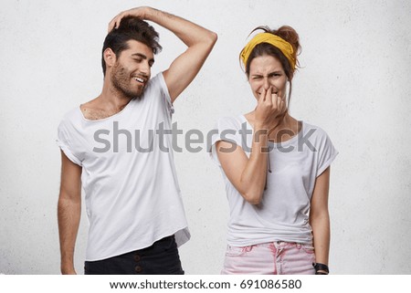 Bad smell and body odor. Picture of fastidious young woman pinching her nose disgusted with terrible sweat, coming out from bearded man's armpit who is standing next to her, raising his arm