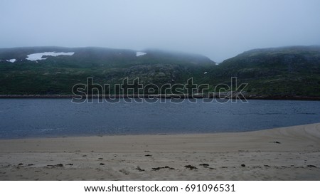 Dense fog covers a big mountains. No trees, just a few fields of grass and sand. Picture is made from another side of little lake.No light comes from a sky. Some snow on a top of hills.No people here.