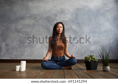 People, meditation and relaxation. Attractive brunette Korean girl wearing top and jeans sitting on wooden floor in half lotus pose, surrounded with candles and plant-pots, meditating with eyes closed