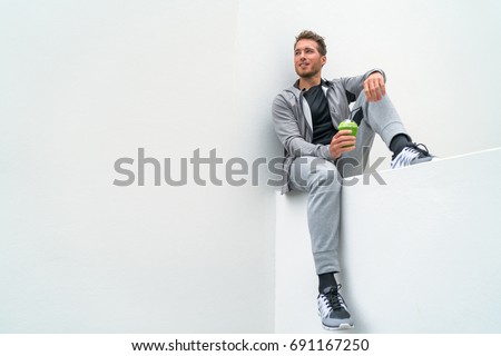 Healthy sport man drinking green smoothie during training at gym in activewear sweatpants outdoor. Athlete sitting relaxing post-workout. Royalty-Free Stock Photo #691167250