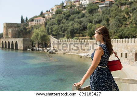 Side view portrait of beautiful woman with strait hair wearing dark blue dress  with white belt and red bag enjoying view picturesque bay Alanya with turquoise water medieval castle wall and shipyard.