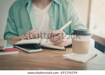 Left-handed Asian freelancer with casual cloths writing on notebook in coffee shop. Outsource worker lifestyle and activity on workday with vintage filter effect