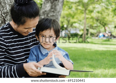 Mother and boy sit under a tree in summer lawn. Happy family playing outdoor Beautiful mother reading a book to her children outside the garden.