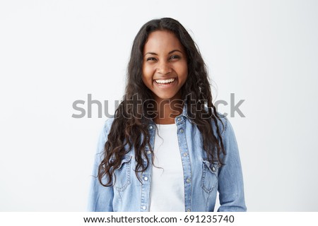 Beautiful dark-skinned female with long brunette hair and broad happy smile wearing denim shirt enjoying good positive news concerning her promotion at work, posing isolated against white blank wall Royalty-Free Stock Photo #691235740