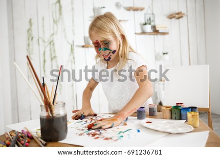 Playful, cute blonde girl having fun by drawing picture with her hands, deeping her palms in different colours and putting them on white sheet of paper. Little laughing girl occupied with drawing in