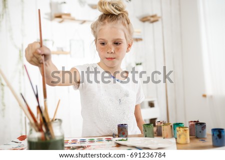 Indoor shot of European cute creative child with hair bun snd blue eyes occupied with drawing. Little blonde girl wearing white t-shirt busy with painting, deeping brush into water, drawing new