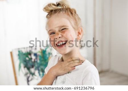 Small European blonde girl with painted face, laughing at camera and squinting in morning light. Creative mood and cheerful atmosphere mixed with shinny look of kid wearing white t-shirt.
