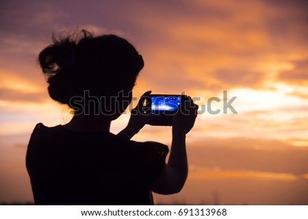 Silhouette of a woman holding a cell phone taking pictures outside during sunset