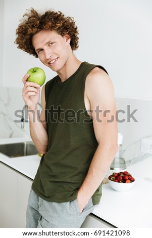 Picture of curly smiling man standing at the kitchen and holding apple. Looking camera.