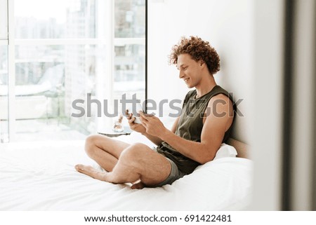 Picture of curly happy man sitting on bed at home using tablet computer. Looking aside.