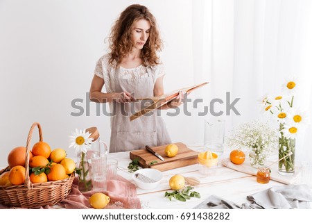 Picture of amazing young woman standing indoors near table with a lot of citruses holding cooking book. Looking aside.