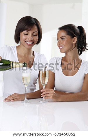 TWO WOMEN WITH DARK BROWN HAIR,IN WHITE INTERIOR, DRESSED IN WHITE DRINKING CHAMPAGNE
