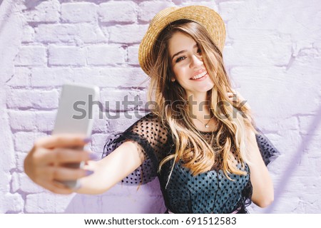 Smiling girl with shiny hair enjoying good weather during walk and making selfie. Outdoor portrait of young laughing woman in romantic outfit taking picture of herself beside white wall.