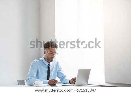 Confident black young male wearing formal clothes, listening to music online on white earphones, resting after hard work at office, sitting in front of opened generic laptop, being always in touch