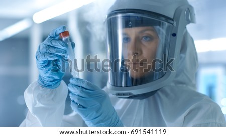 Medical Virology Research Scientist Works in a Hazmat Suit with Mask, Inspects Test Tube with Isolated Virus String from Refrigerator Box. She Works in a Sterile High Tech Laboratory Facility. Royalty-Free Stock Photo #691541119