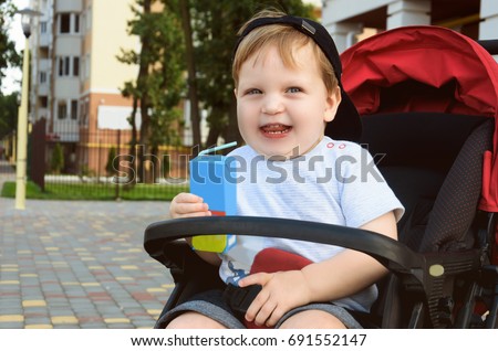 Little boy is sitting in a pram and drinking juice from a packet through a drinking straw and laughing