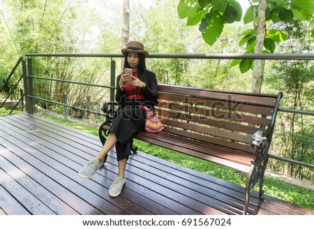 Beautiful asian woman sit on wood chair use phone in the park. Tourist sit on wooden bench send picture on facebook in flower garden., copy space for text.