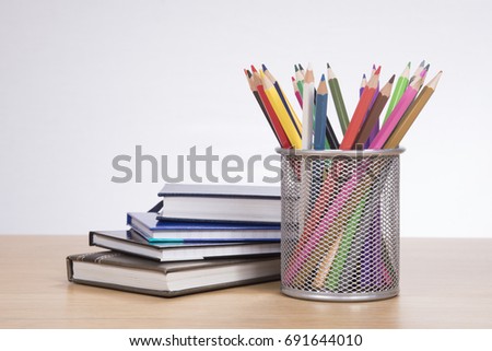 Collection of brightly colored pencil crayons in a metal container on a wooden desk alongside a pile of school books or office journals