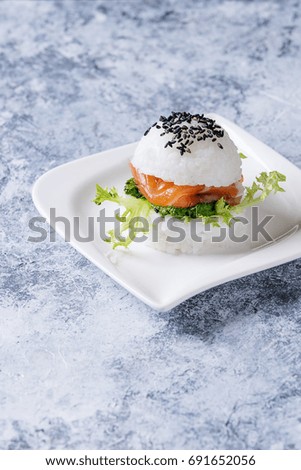 Mini rice sushi burger with smoked salmon, green salad and sauces, black sesame served on white square plate over gray concrete background. Modern healthy food