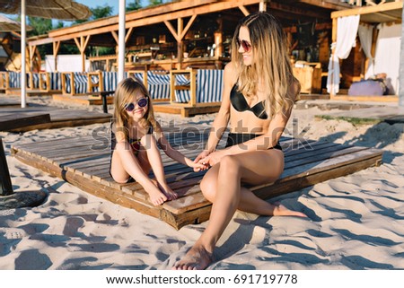 Picture of  mother with little beautiful daughter dressed in black swim suits on the summer beach. They are looking on each other with funny real emotions. Summer day. Background sandy beach.
