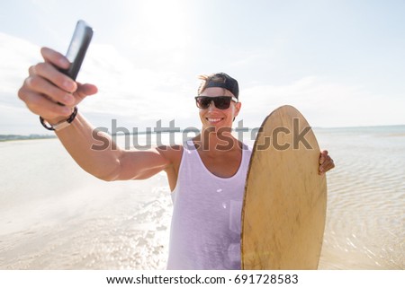 skimboarding, water sport and people concept - happy smiling young man with skimboard and smartphone taking selfie on summer beach