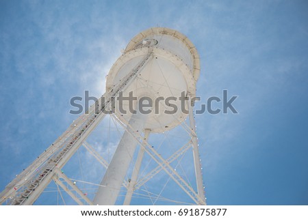 Photo looking up at a white water tower with a bright blue sky and back-lit by the sun.