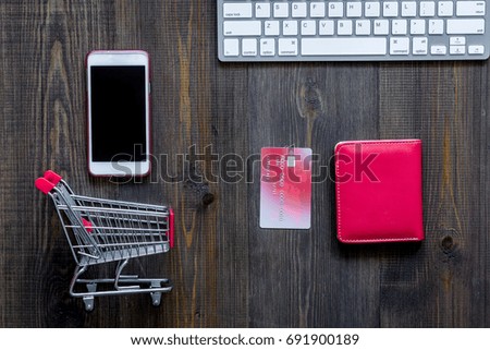 Online shopping concept. Shopping trolley near bank card, keyboard, phone on dark wooden background top view copyspace