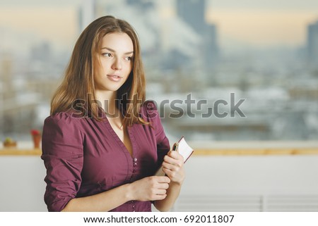 Portrait of pretty thoughtful young woman with book on blurry office interior with city view background. Education concept 