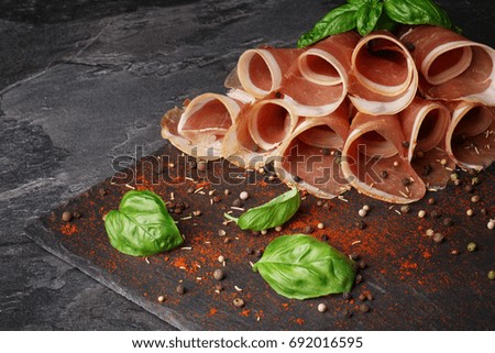 Beautiful picture of tasty twisted meat slices on a brown cutting board. Beautiful red ham, green basil leaves, red spices on a dark gray stone background. Delicious appetizers for beer. Copy space.