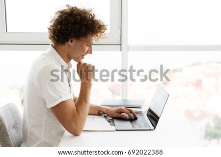 Picture of handsome concentrated serious man sitting at the table near window indoors using laptop computer. Looking aside.