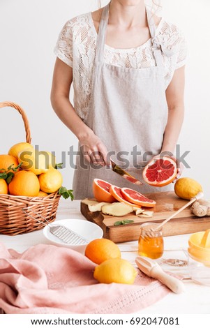 Cropped picture of young woman standing indoors near table with a lot of citruses and cut the grapefruit.