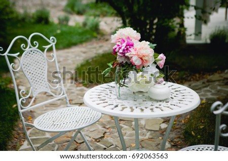 A beautiful forged coffee table with white chairs stands in the garden among green bushes and trees. Decoration from flowers. Location for her morning photo session