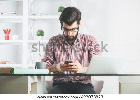 Portrait of attractive young businessman using mobile phone while sitting at office desk with laptop and other items. Leisure, break, communication and technology concept