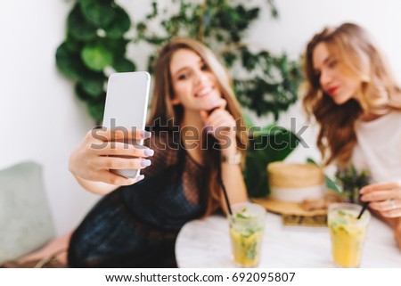 Gorgeous blonde young woman in stylish attire taking picture of herself on plant background while spending time with friend in cafe. Indoor portrait of two stylish girls enjoying juice in restaurant.