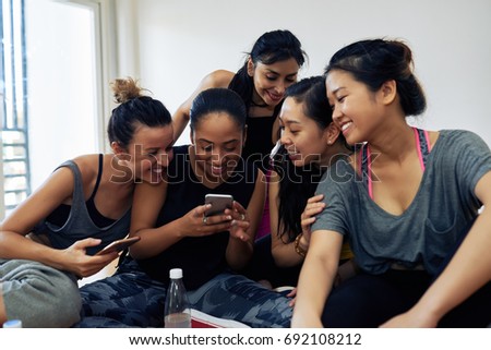 Multi-ethnic group of friends in sportswear gathered together in fitness studio after intensive training and looking through photos on smartphone