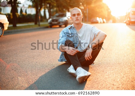 Handsome young youth man in stylish fashionable denim clothes sits on asphalt road at sunset