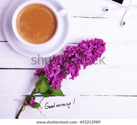 Black coffee in a white cup with a saucer, near a bud of purple lilac with a paper tag and the inscription good morning, top view