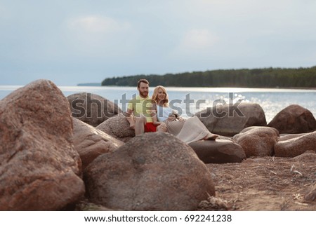 A man in shorts and a t-shirt and a girl in a summer dress enjoying each other. Beautiful couple sitting on a rock on the beach, watching the sunset. They wear casual clothes