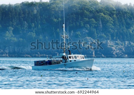 A white lobster boat with lobster traps piled on the back of the boat, heads out to sea to set traps. One of the porcupine islands with its tall green trees is in the background
