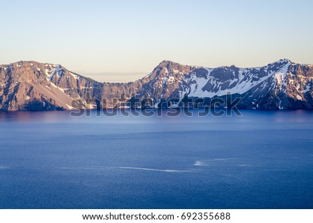 Landscape of Crater Lake, the deepest lake in USA. Deep blue water in the foreground, followed by snowy mountains and sky dividing the picture in three parallel layers in morning light.