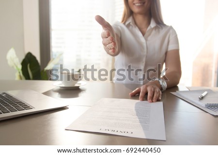 Smiling businesswoman proposes partnership, gives hand at camera for handshake, offers to sign contract, makes business proposition, good deal proposal, ready to conclude agreement at desk, close up