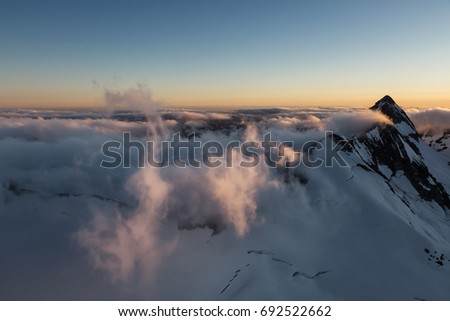 Beautiful aerial landscape view of the snow covered mountains with glowing clouds from the sunlight during sunset. Picture taken from an airplane near Squamish, North of Vancouver, BC, Canada.
