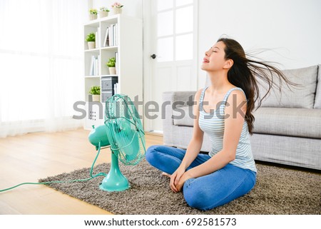 happy elegant woman blowing electric fan in summer day and sitting on living room floor enjoying cool wind to refreshing. Royalty-Free Stock Photo #692581573