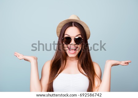 Young cute brunette toothy  lady in summer outfit and trendy summer tourist`s hat is standing on a light blue background, with hands on sides, holding the copy space