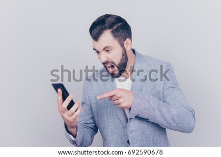 Going crazy and insane, psycho and agression. Close up portrait of shouting stressed young brunet bearded entrepreneur, pointing at his phone, on pure light background, wearing casual smart