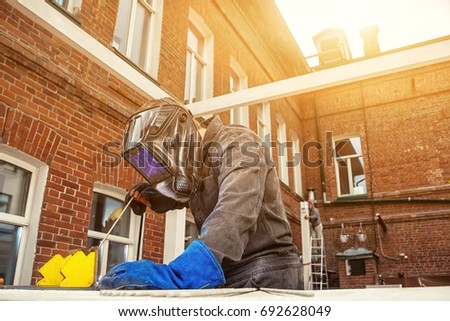 A strong man welder in a construction black overall and welding brush welds a metal welding machine, in the background an ancient wooden building and the sun shines brightly